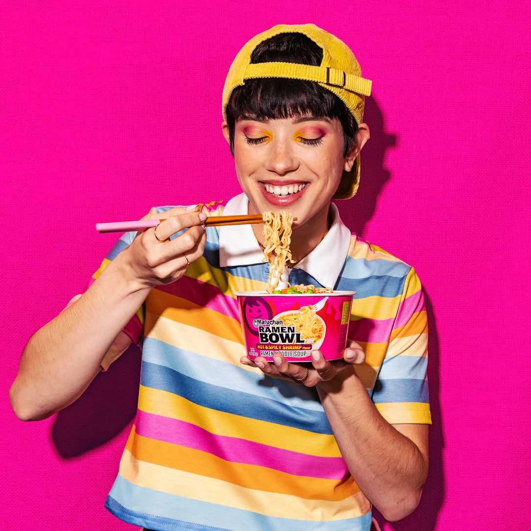Person smiling while eating noodles with chopsticks from a Maruchan ramen bowl