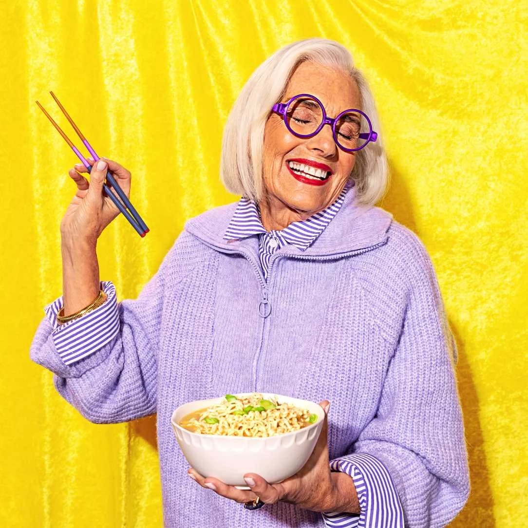 Older woman smiling while holding a bowl of ramen noodles and holding a pair of chopsticks in the other hand