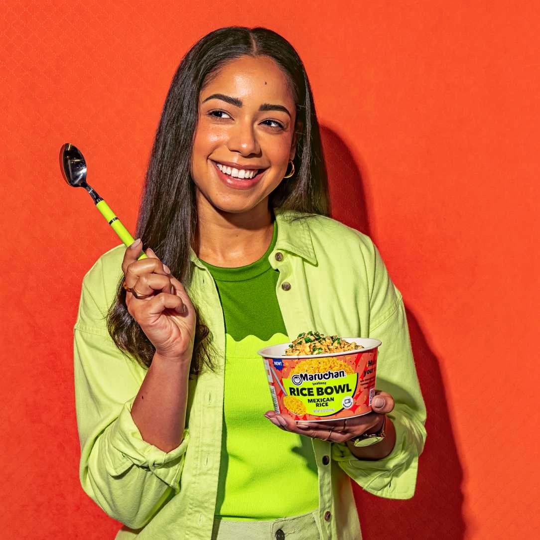 Woman smiling while holding a spoon in one hand and Maruchan rice bowl in her other hand