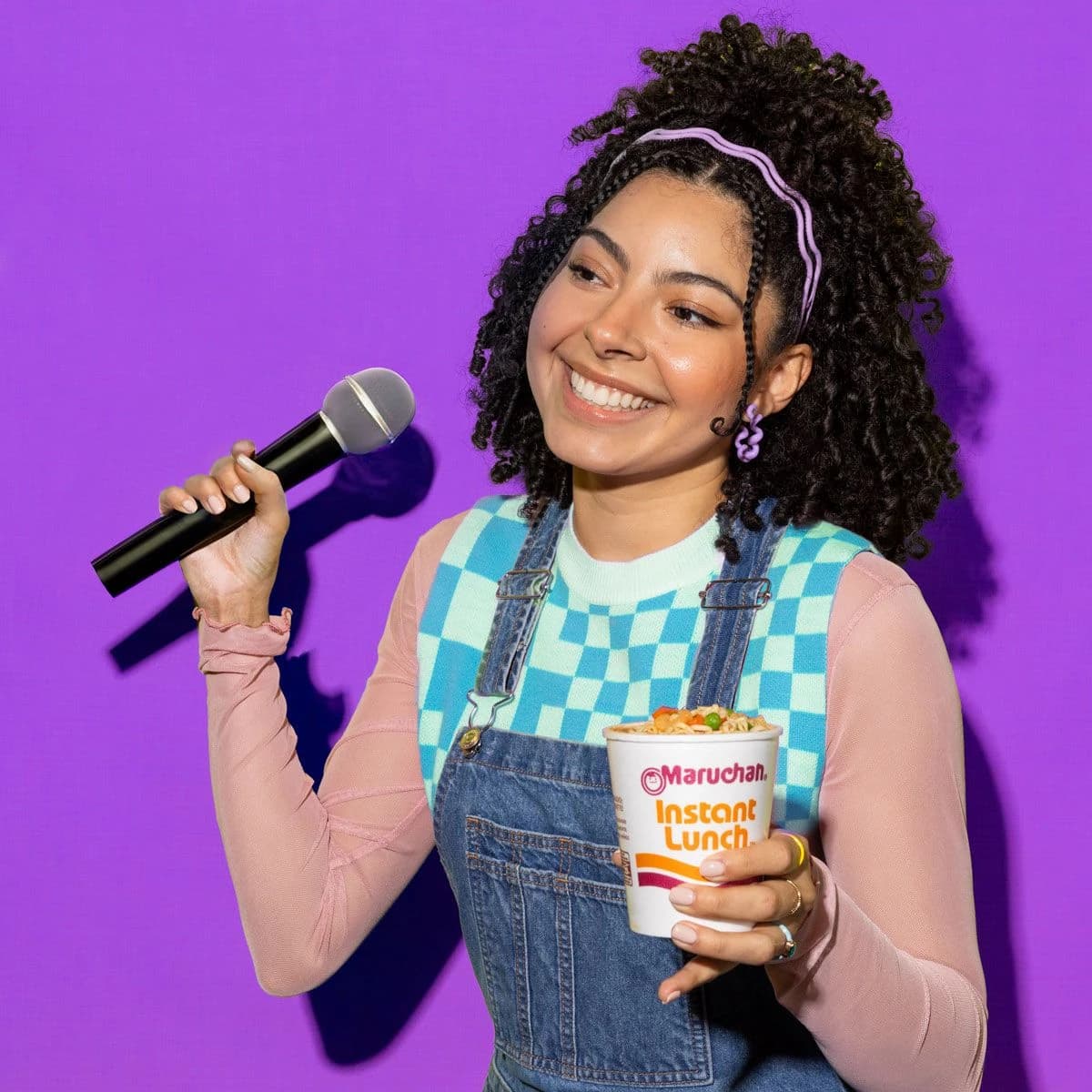 Woman smiling and holding a microphone in one hand and a cup of instant lunch noodles in her other hand