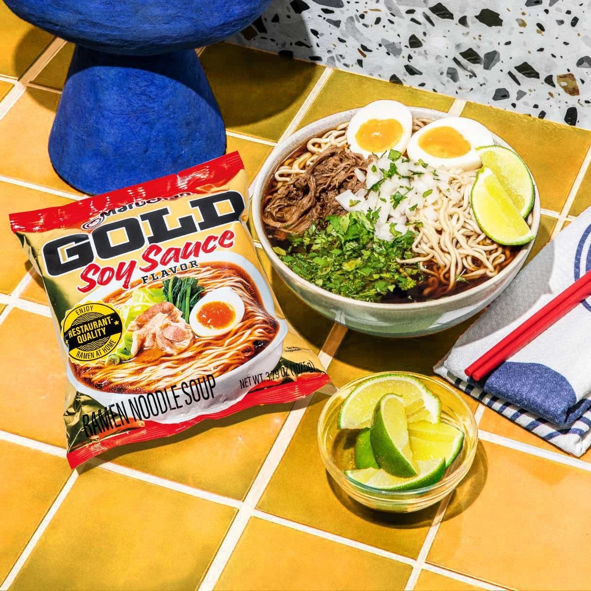 A packet of gold soy sauce flavor ramen next to a prepared bowl of ramen with birria beef and eggs next to a bowl of limes.
