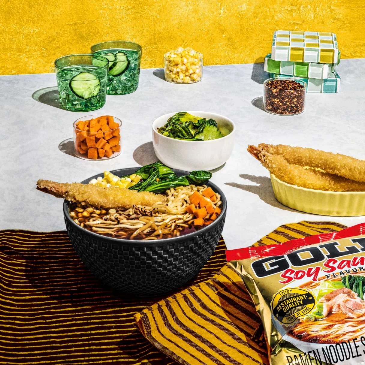 A bowl of gold soy sauce ramen with bowls of vegetables and tempura against a yellow background.