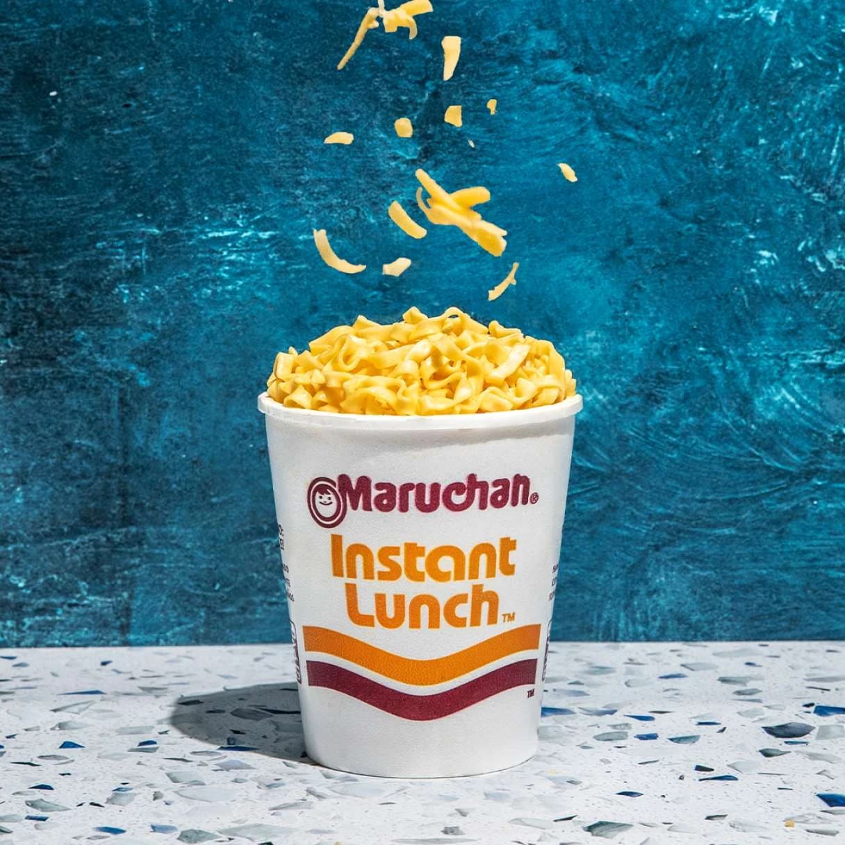 Instant lunch cheddar cheese flavor ramen cup on a table with cheese being sprinkled on top